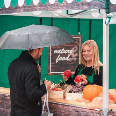 La venditrice nella tenda del mercato tiene in mano diversi frutti. Il cliente è in piedi davanti alla tenda di vendita al banco.