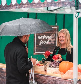 The saleswoman in the market tent holds various fruits in her hand. The customer is standing in front of the sales tent at the counter.