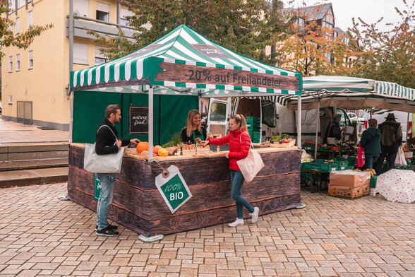 Der Marktstand ist vollflächig bedruckt und kommt auf dem Wochenmarkt und Bauernmarkt zum Einsatz. 2 Kunden sehen sich die Produkte an.