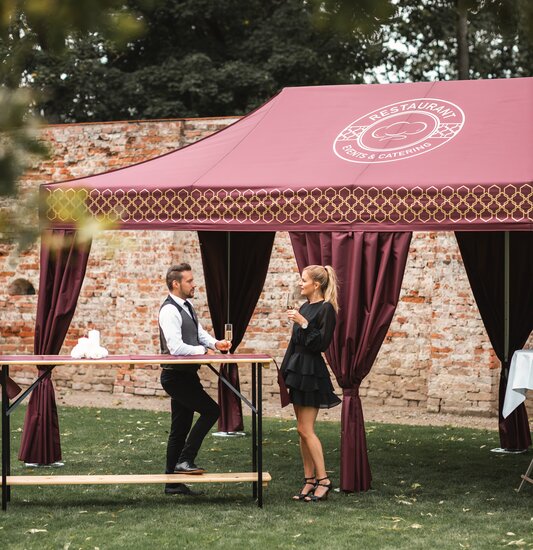 Im großen Garten steht das bordeaux-farbene Cateringzelt. Die Frau und der Mann haben ein Sektglas in der Hand und stehen an einem Stehtisch.