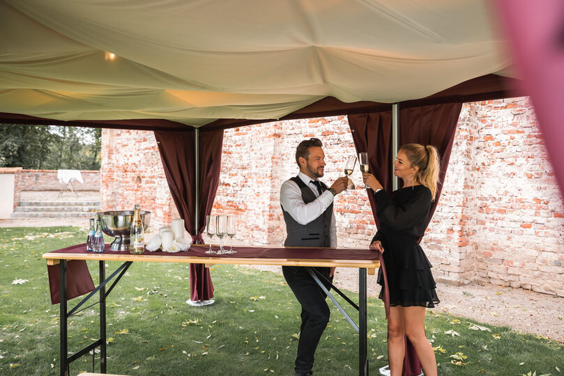 Under the elegant folding gazebo with a sky, 2 people stand with a glass of prosecco in their hands. 