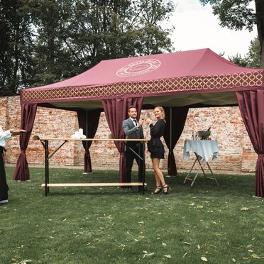 Il gazebo catering color bordeaux si trova in un prato. Una coppia è in piedi davanti ad essa con un bicchiere di prosecco in mano. La cameriera porta degli stuzzichini su un vassoio.