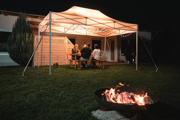 Das 4,5x3 m Gartenzelt steht im Dunkeln im Garten. Darunter sitzen die 4 Gäste. Vor dem Zelt ist noch eine Feuerstelle.