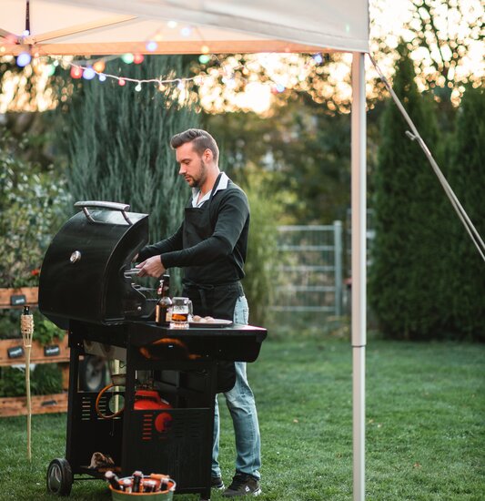 Der Mann steht mit seinem Grill unter einem ecru-farbenen Faltpavillon im Garten. Der Faltpavillon ist dekoriert mit Lichterketten und gesichert mit Spanngurten.