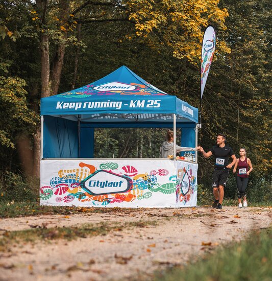 Blauer 3x3m Eventzelt mit individuellem Citylauf-Druck, mit individueller Fahne, geschlossener Seitenwand und halbhoher Seitenwand mit Theke als Verpflegungsstand bei einem Stadtlauf