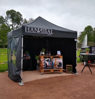 Gazebo 3x3 nero con 2 pareti laterali nere chiuse e una parete laterale con finestra panoramica personalizzato con logo Hannibal Gin utilizzato per la vendita di gin su una stradina.